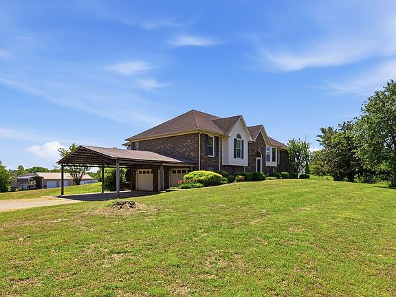 Eastern approach to 1474 Hardison Rd, the open carport and attached garage set against the brick facade as the lawn sweeps down through five fenced acres